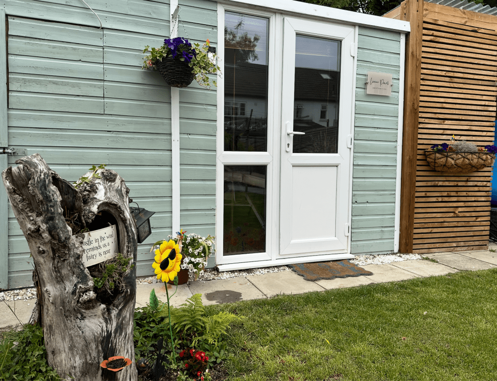 Exterior view of a home aesthetics clinic featuring a white door, wooden signage, and decorative plants.